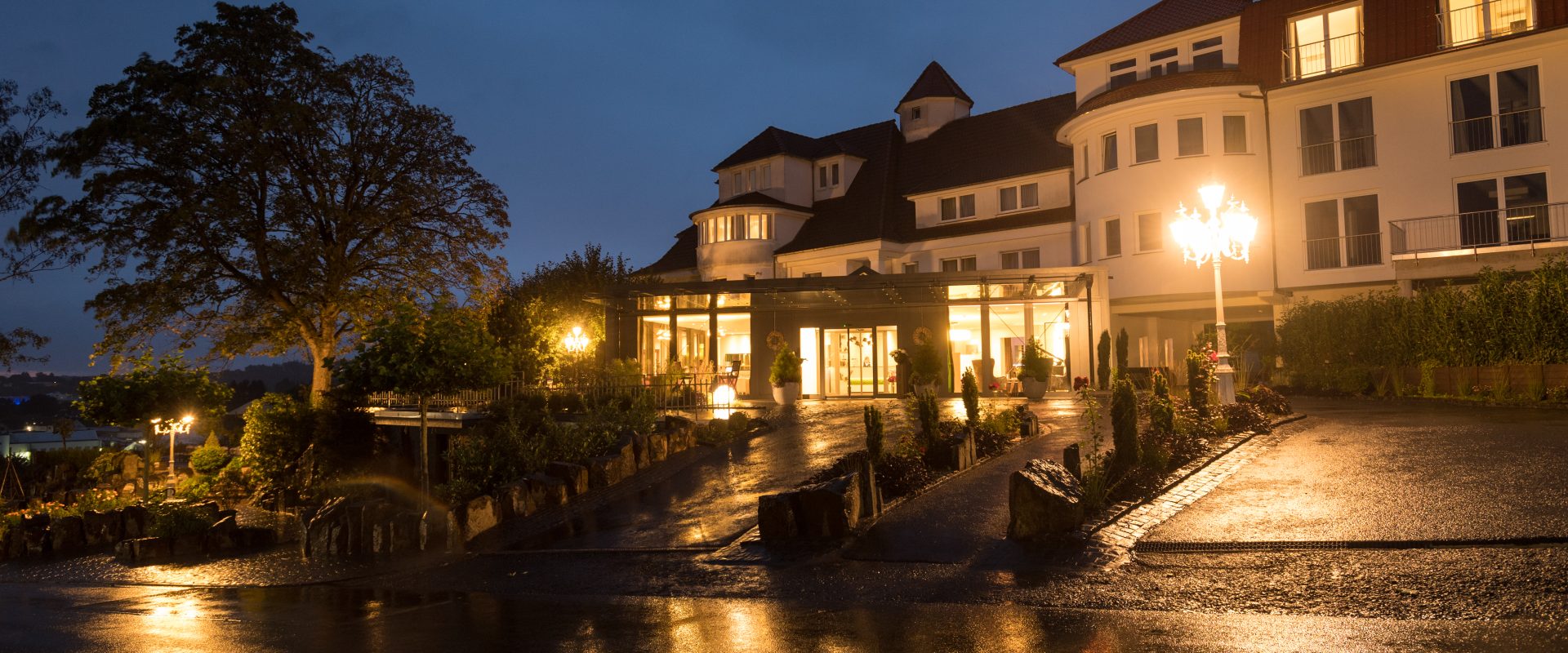 In der Abenddämmerung steht ein warm beleuchtetes Hotel mit rotem Dach, dessen Lichter sich nach dem Regen auf der nassen Auffahrt spiegeln.