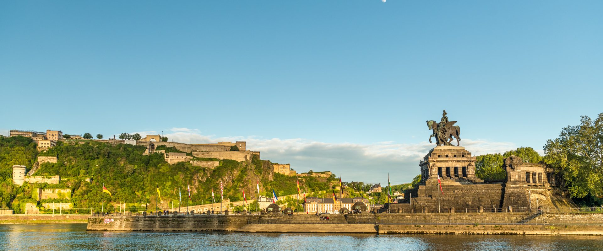Blick auf einen großen Fluss, eine historische Festung auf einem Hügel und ein Reiterstandbild unter einem klaren blauen Himmel mit sichtbarem Mond.