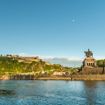Blick auf einen großen Fluss, eine historische Festung auf einem Hügel und ein Reiterstandbild unter einem klaren blauen Himmel mit sichtbarem Mond.