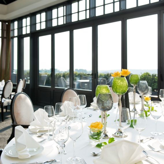 Eleganter runder Esstisch mit Glaswaren, weißen Servietten und gelben Blumengestecken an großen, sonnendurchfluteten Fenstern.