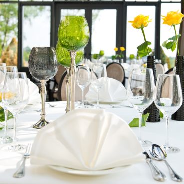 Elegante Tischdekoration mit gefalteten Servietten, Glaswaren und gelben Rosen in Vasen auf einer weißen Tischdecke in einem hellen Esszimmer.