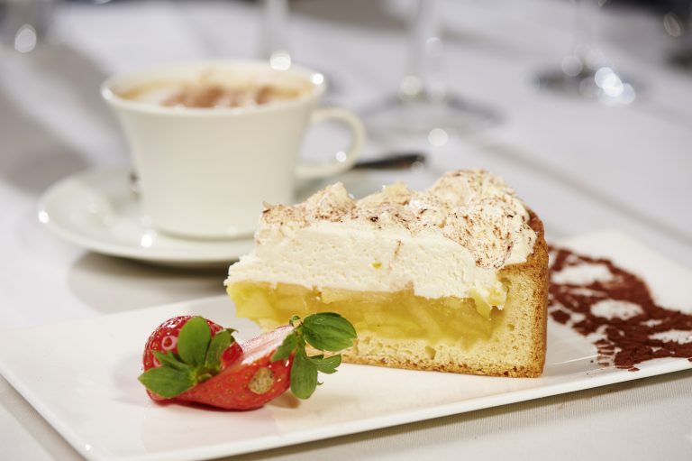 Ein Stück Kuchen mit Schlagsahne, garniert mit einer Erdbeere, serviert auf einem Teller neben einer Tasse Kaffee.