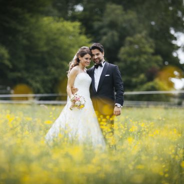 Eine Braut und ein Bräutigam stehen lächelnd zusammen auf einem Feld mit gelben Wildblumen und grünen Bäumen im Hintergrund.