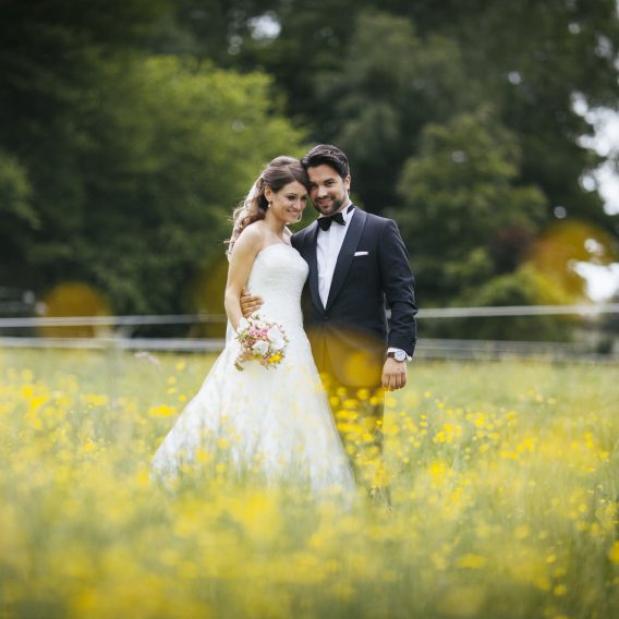 Eine Braut und ein Bräutigam stehen lächelnd zusammen auf einem Feld mit gelben Wildblumen und grünen Bäumen im Hintergrund.