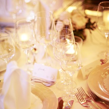 Elegant gedeckter Esstisch mit Tellern, Besteck, Weingläsern, Servietten, Namenskarten und sanftem Kerzenlicht neben weißen Blumen.
