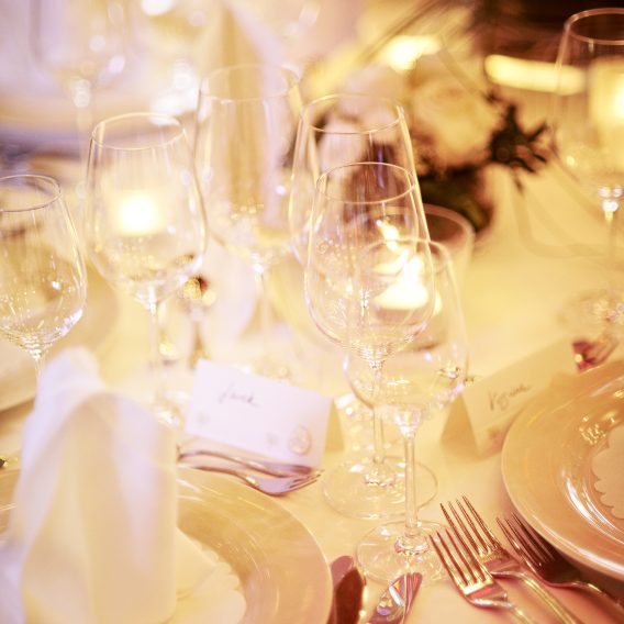 Elegant gedeckter Esstisch mit Tellern, Besteck, Weingläsern, Servietten, Namenskarten und sanftem Kerzenlicht neben weißen Blumen.