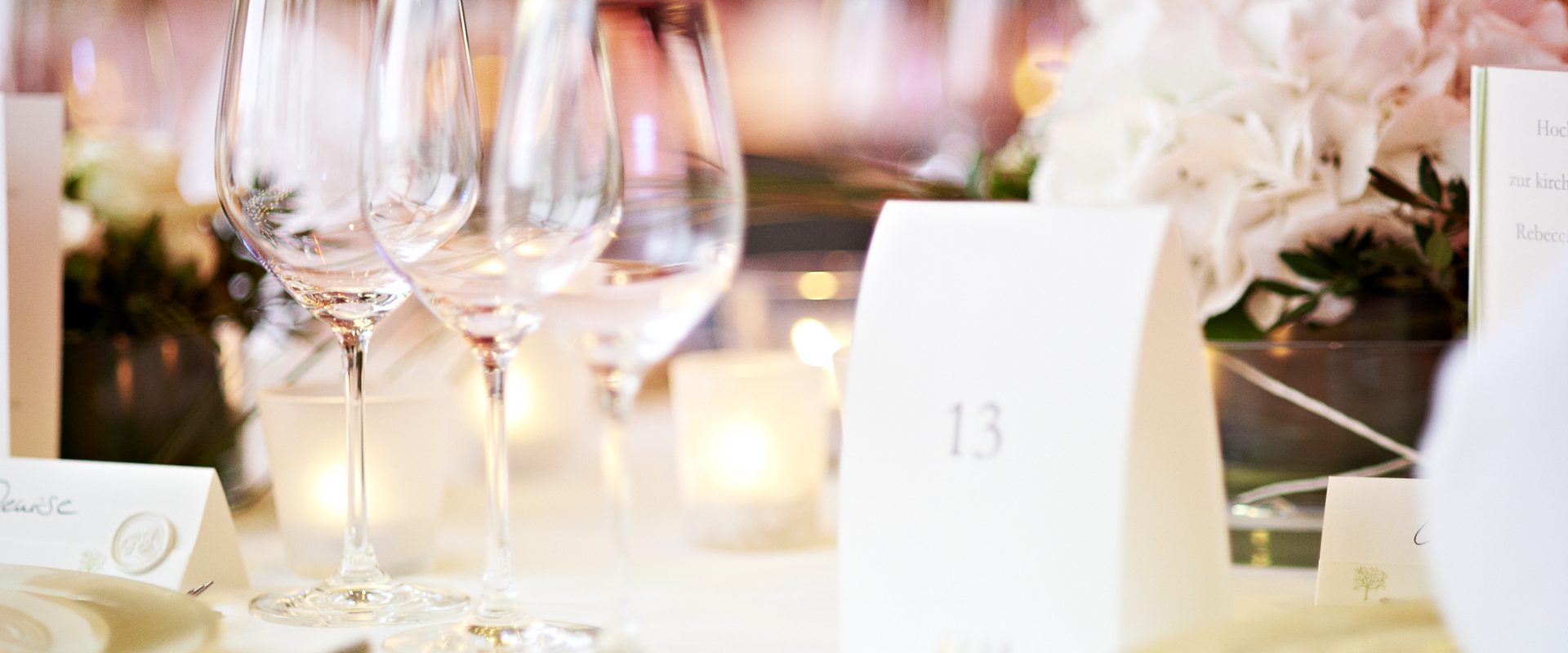 Elegante Tischdekoration mit Weingläsern, weißen Blumen und einer Tischnummernkarte, sanft beleuchtet für eine formelle Veranstaltung oder Feier.
