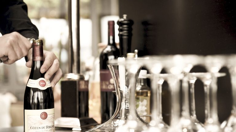 Eine Person öffnet eine Flasche Rotwein. In der Nähe stehen Weingläser, eine Karaffe und Flaschen auf einem Tisch in einer sanft beleuchteten Umgebung.