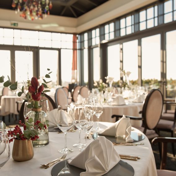 Elegantes, sonnendurchflutetes Restaurant mit runden Tischen, die mit weißer Tischwäsche, gefalteten Servietten, Glaswaren und Blumengestecken gedeckt sind.