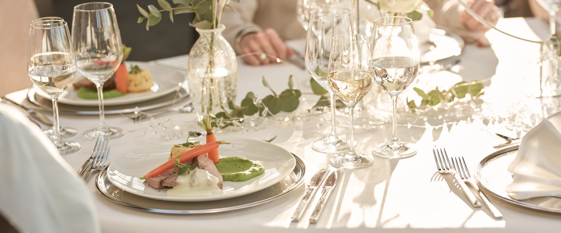 Elegant gedeckter Tisch mit weißen Rosen und Weingläsern; Gourmet-Essen auf Tellern in sanftem Tageslicht, Menschen speisen gemeinsam.