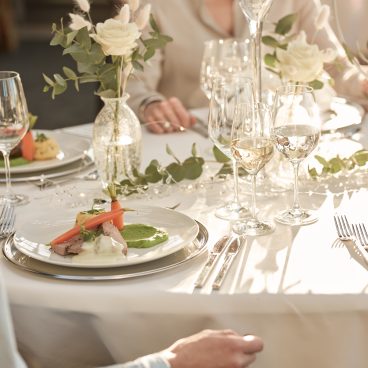 Elegant gedeckter Tisch mit weißen Rosen und Weingläsern; Gourmet-Essen auf Tellern in sanftem Tageslicht, Menschen speisen gemeinsam.