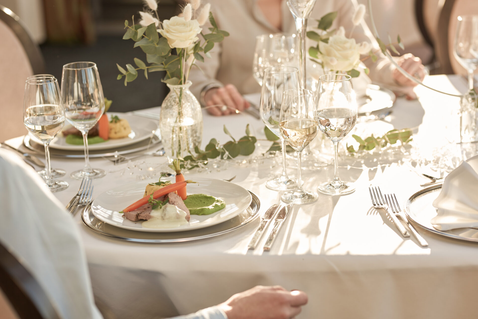 Elegant gedeckter Tisch mit weißen Rosen und Weingläsern; Gourmet-Essen auf Tellern in sanftem Tageslicht, Menschen speisen gemeinsam.