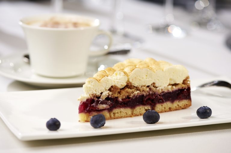 Ein Stück Beerenkuchen mit Sahnehaube wird auf einem weißen Teller mit frischen Blaubeeren neben einer Tasse Kaffee serviert.