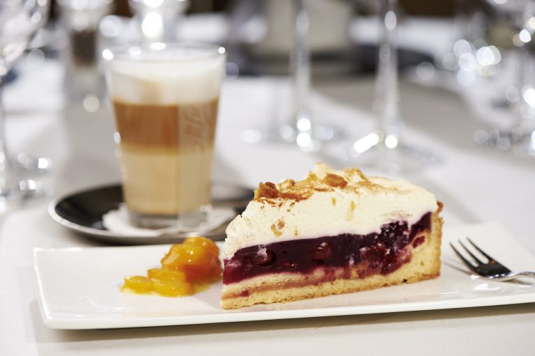 Ein Stück Beerenkuchen mit Schlagsahne, Mangostücken und einem Glas Schichtkaffee auf einem weißen Teller mit einer Gabel.