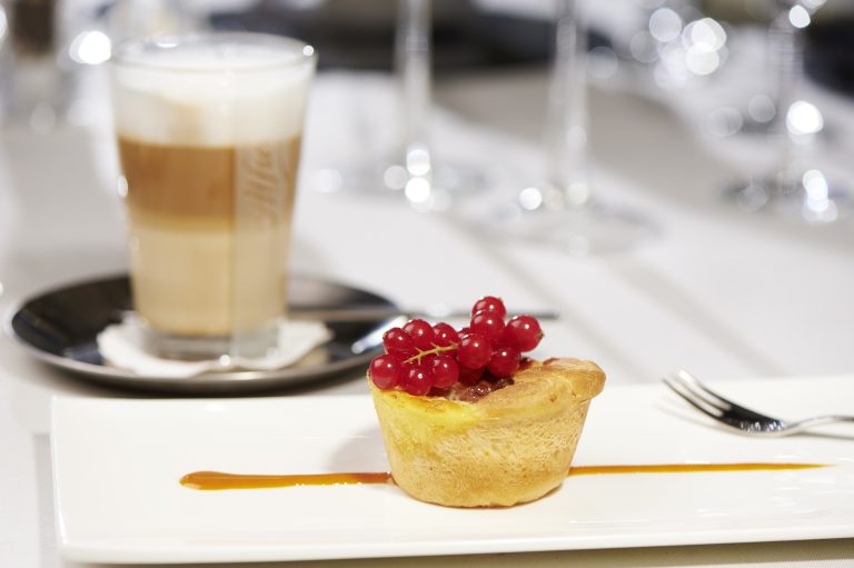 Eine Desserttorte mit roten Johannisbeeren auf einem weißen Teller, daneben eine Gabel und ein Glas geschichteter Latte.