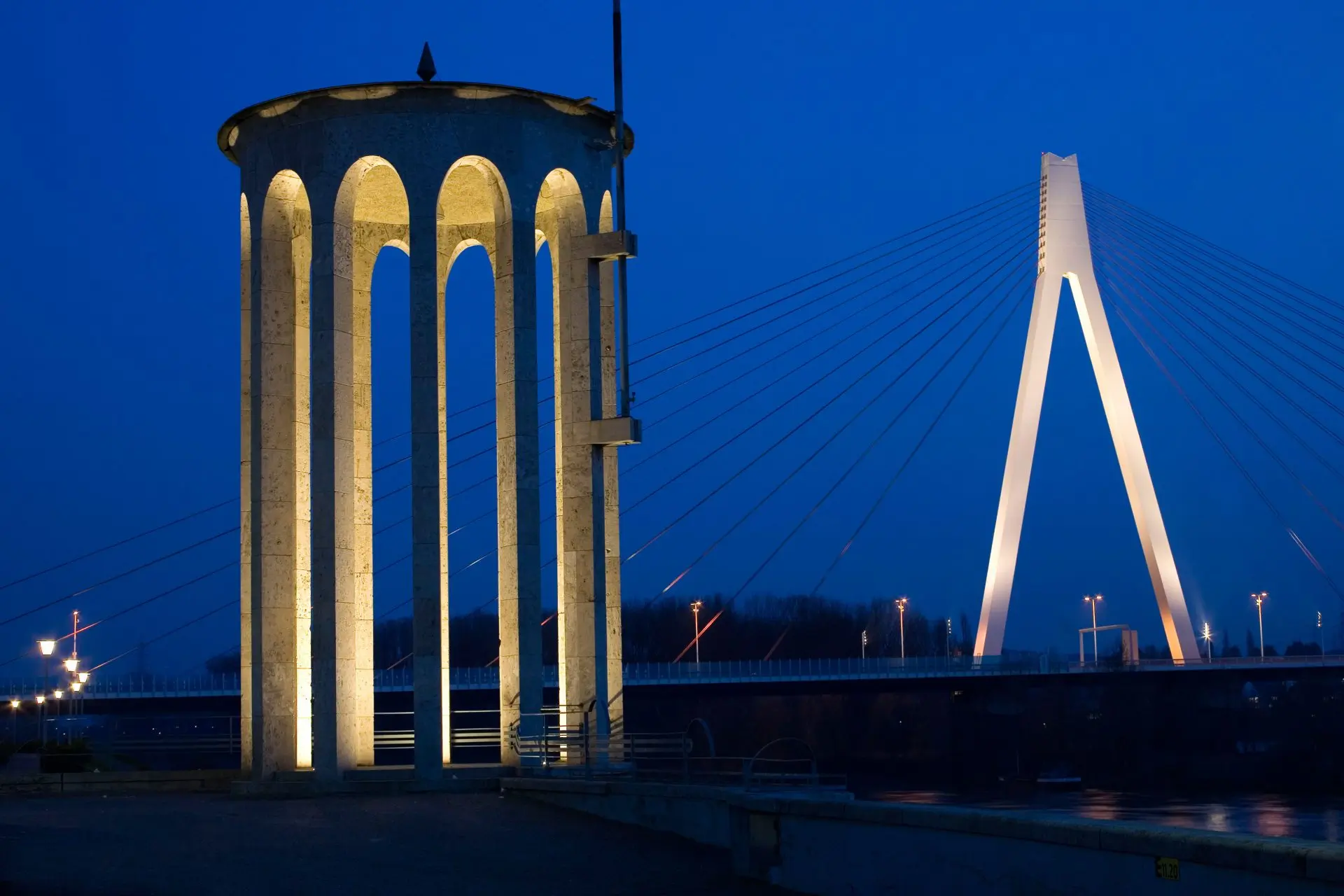 Beleuchtetes Bogendenkmal und moderne Schrägseilbrücke bei Nacht, beide vor einem tiefblauen Himmel beleuchtet.
