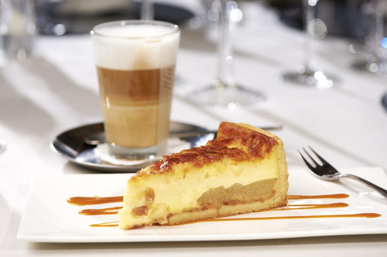 Ein Stück Käsekuchen mit Karamellüberzug auf einem Teller, neben einem Glas geschichtetem Latte auf einem Tisch.