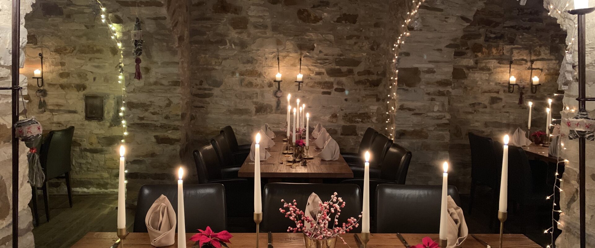 Ein gemütliches Esszimmer mit Steinwänden, Kerzen, gefalteten Servietten, Tannenzapfen und Weihnachtsdekorationen auf Holztischen.