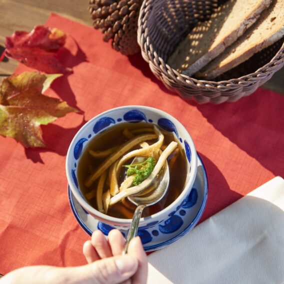 Eine Hand hält einen Löffel über einer Schüssel Nudelsuppe, auf dem Tisch liegen Brot, Herbstblätter und ein Tannenzapfen auf einem roten Tuch.