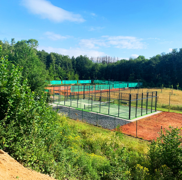 Tennis- und Padelplätze im Freien, umgeben von Grün und Bäumen unter einem strahlend blauen Himmel.