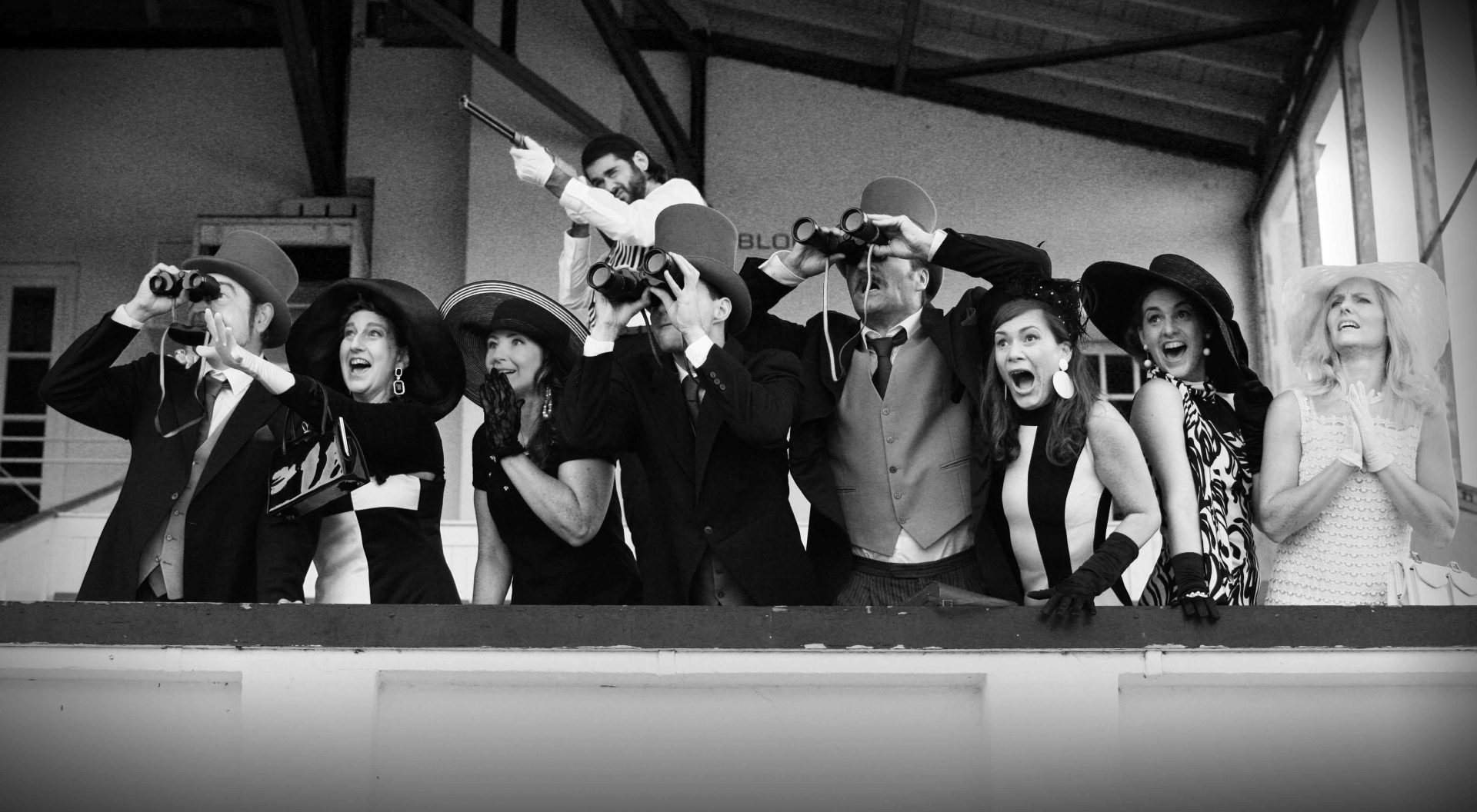 Eine Gruppe von Menschen in Vintage-Kleidung jubelt und beobachtet gespannt ein Pferderennen, manche mit Ferngläsern.