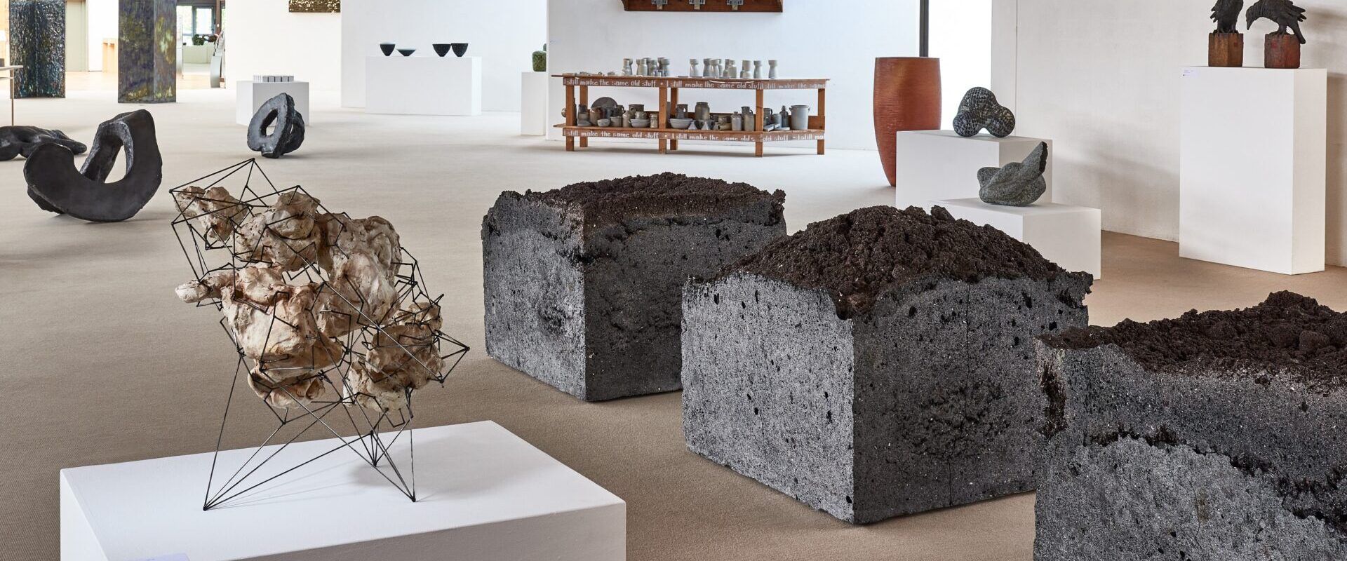 Galerie für zeitgenössische Kunst mit abstrakten Skulpturen, Erdwürfelinstallationen und Töpferwaren, die in einem geräumigen Raum auf Podesten und Tischen ausgestellt sind.