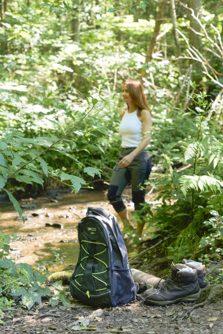 Eine Frau steht barfuß in einem Bach, am Rand ein Wanderrucksack und Wanderstiefel