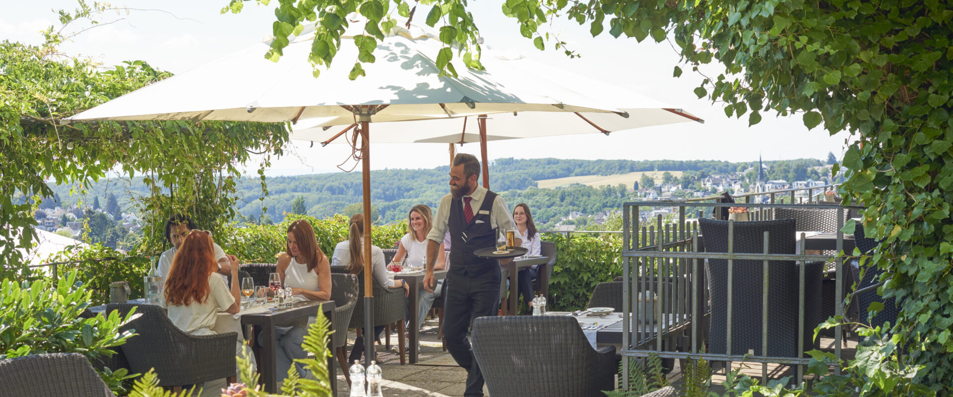 Mehrere Menschen sitzen an Tischen auf der Terrasse, sprechen, essen und trinken, ein Kellner läuft vorbei