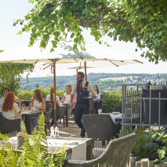 Mehrere Menschen sitzen an Tischen auf der Terrasse, sprechen, essen und trinken, ein Kellner läuft vorbei