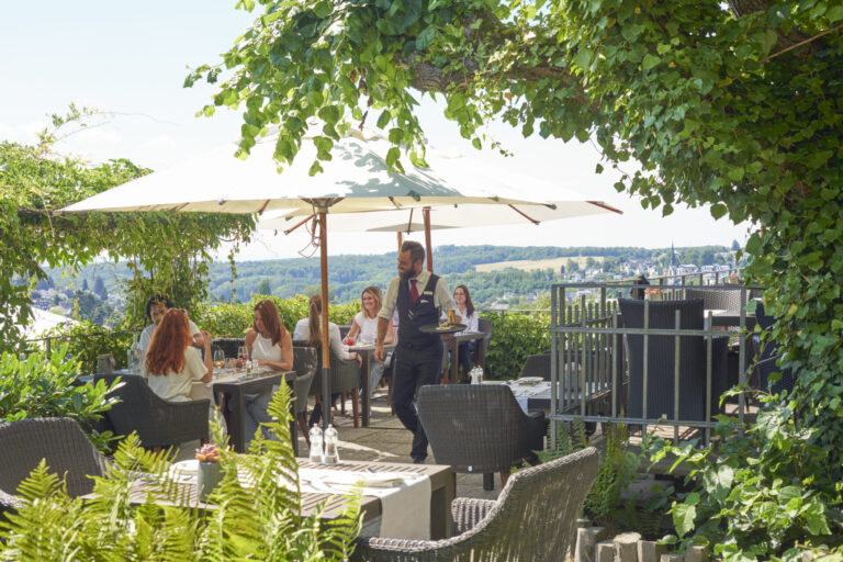 Mehrere Menschen sitzen an Tischen auf der Terrasse, sprechen, essen und trinken, ein Kellner läuft vorbei