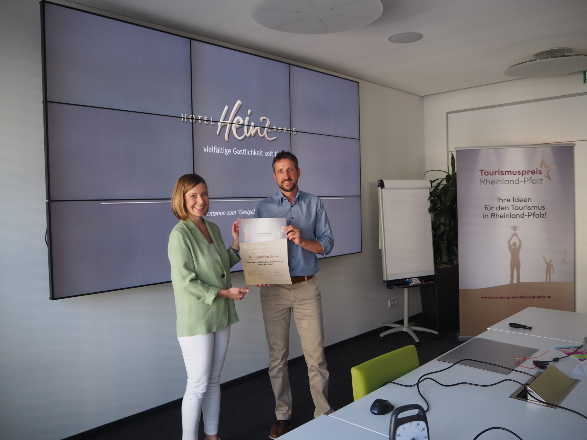 Eine Frau und ein Mann stehen zusammen und halten ein Zertifikat vor einem Bildschirm und einem Banner mit einer Tourismusauszeichnung in einem Büro.