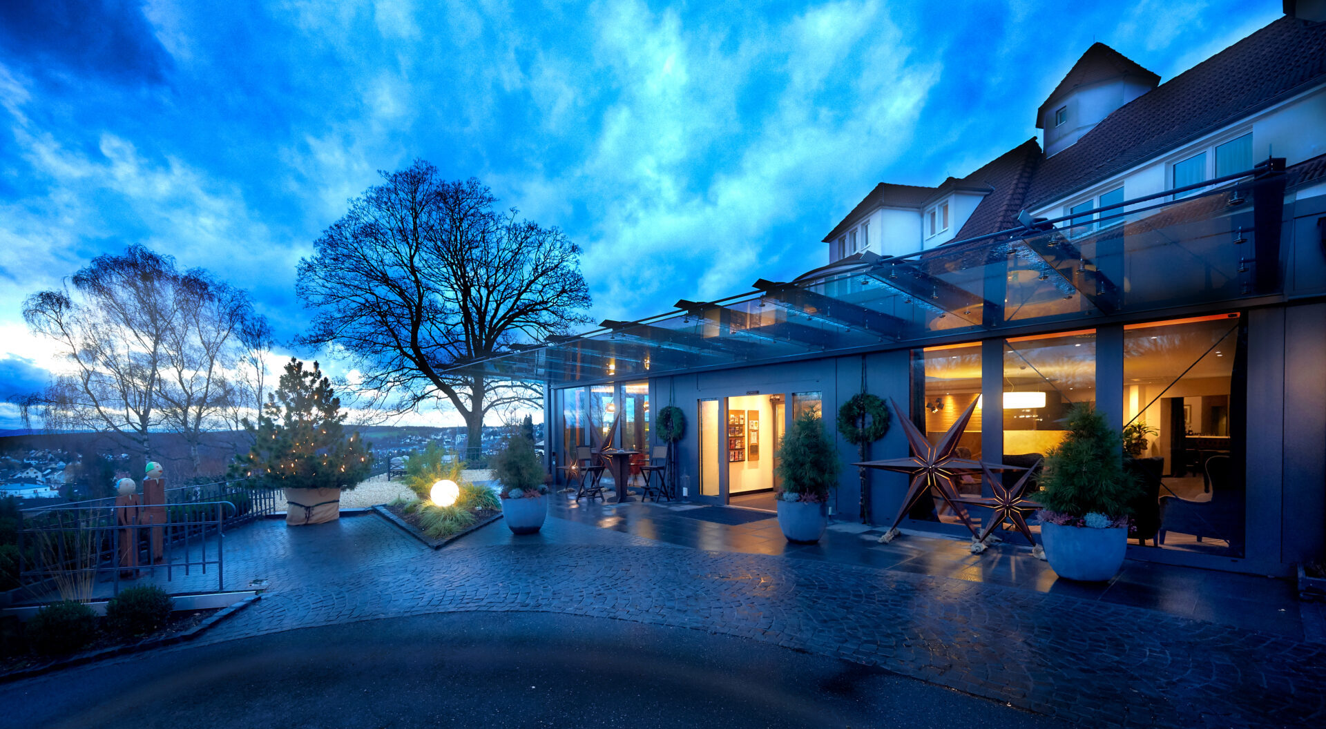 Modernes Hotel mit Glasfenstern in der Abenddämmerung, warmer Innenbeleuchtung, Topfpflanzen und Blick auf Bäume und eine Stadt unter einem dramatisch blauen Himmel.