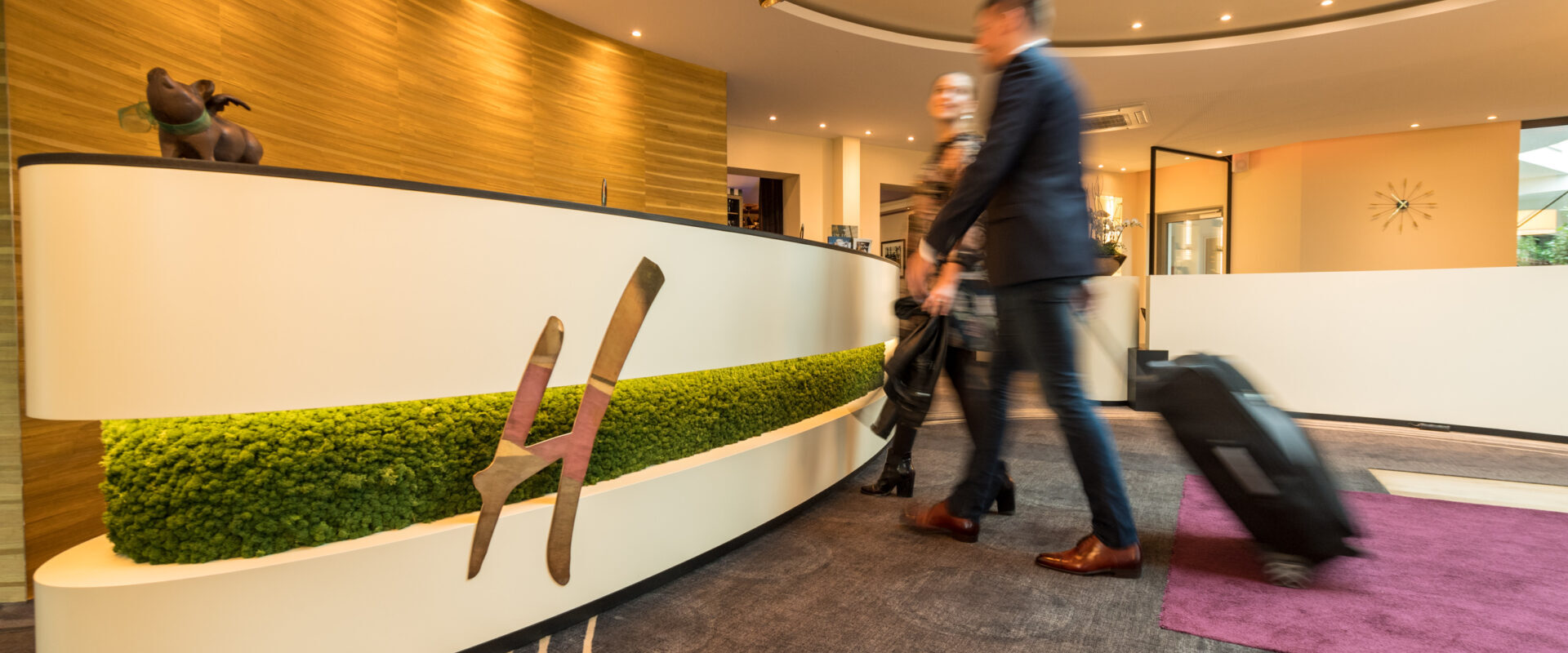 Zwei Personen mit Koffern gehen an einer modernen Hotelrezeption mit grünem Akzent und einer dekorativen Hundestatue vorbei.