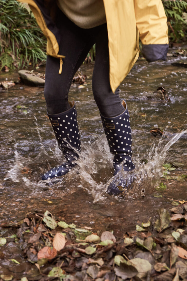 Person in gelbem Regenmantel und gepunkteten Stiefeln planscht in einem flachen, steinigen Bach, umgeben von gefallenen Blättern.