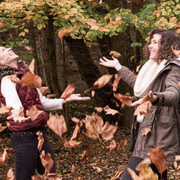 Zwei Frauen werfen freudig Herbstblätter in die Luft, während sie in einem Wald stehen, der von Bäumen mit buntem Laub umgeben ist.