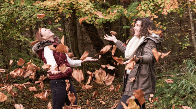 Zwei Frauen werfen freudig Herbstblätter in die Luft, während sie in einem Wald stehen, der von Bäumen mit buntem Laub umgeben ist.