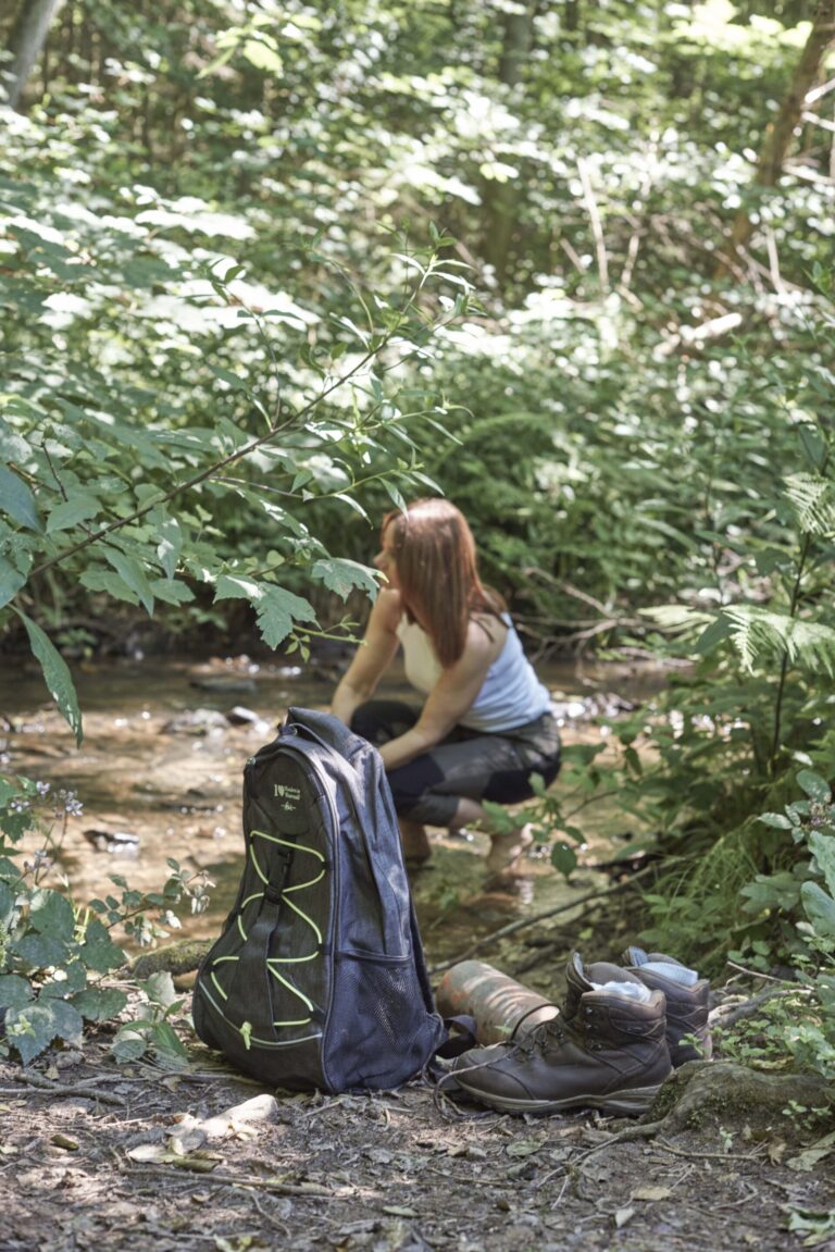 Eine Frau hockt an einem Waldbach, im Vordergrund liegen ein Rucksack und Wanderschuhe auf dem Boden.