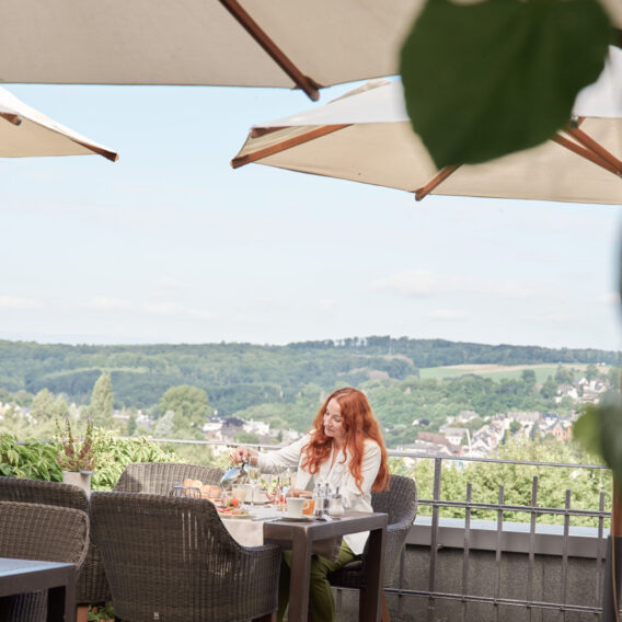 Eine Frau mit langen roten Haaren speist allein an einem Tisch auf einer Terrasse im Freien, umgeben von Grün und mit Blick auf eine malerische Hügellandschaft.
