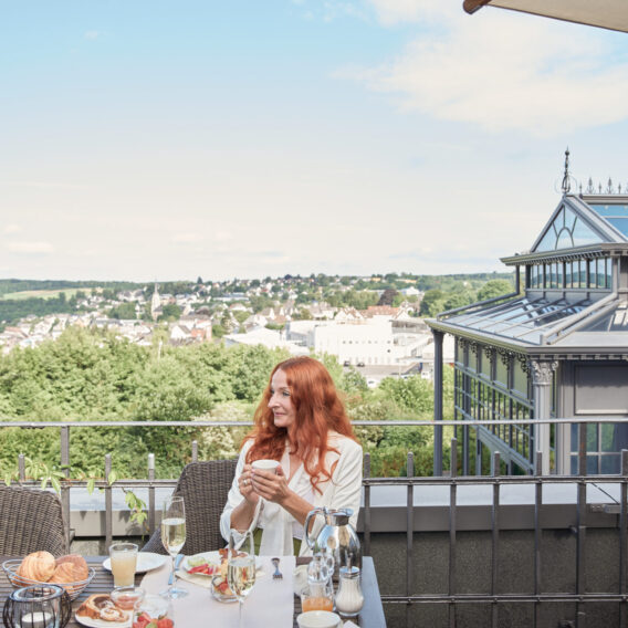 Eine Frau mit langen roten Haaren genießt ihr Frühstück an einem Tisch im Freien mit einem malerischen Blick auf die Stadt und das Grüne im Hintergrund.