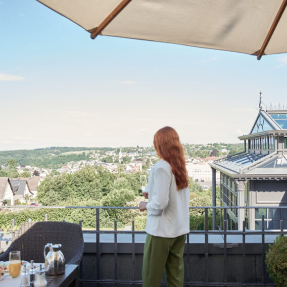 Eine Frau mit langen roten Haaren steht auf einer Terrasse unter einem Sonnenschirm und blickt auf eine malerische Stadt und eine grüne Landschaft.