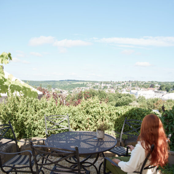 Eine Frau mit langen roten Haaren sitzt an einem Terrassentisch und blickt auf eine malerische, sonnenbeschienene Landschaft mit viel Grün und Gebäuden in der Ferne.