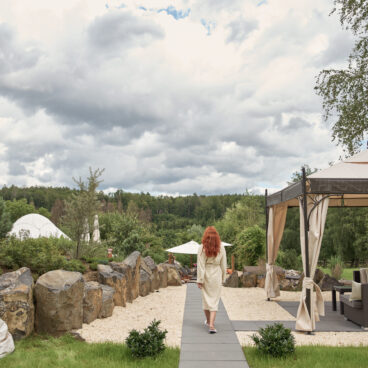 Eine Frau mit roten Haaren geht unter einem bewölkten Himmel auf einem Steinpfad durch einen Garten mit Felsen, Zelten und einem Pavillon.
