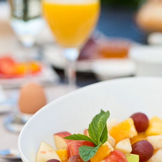 Eine Schüssel mit frischem Obstsalat mit Trauben und Minze, im Hintergrund sind Orangensaft und andere Frühstückszutaten verschwommen.