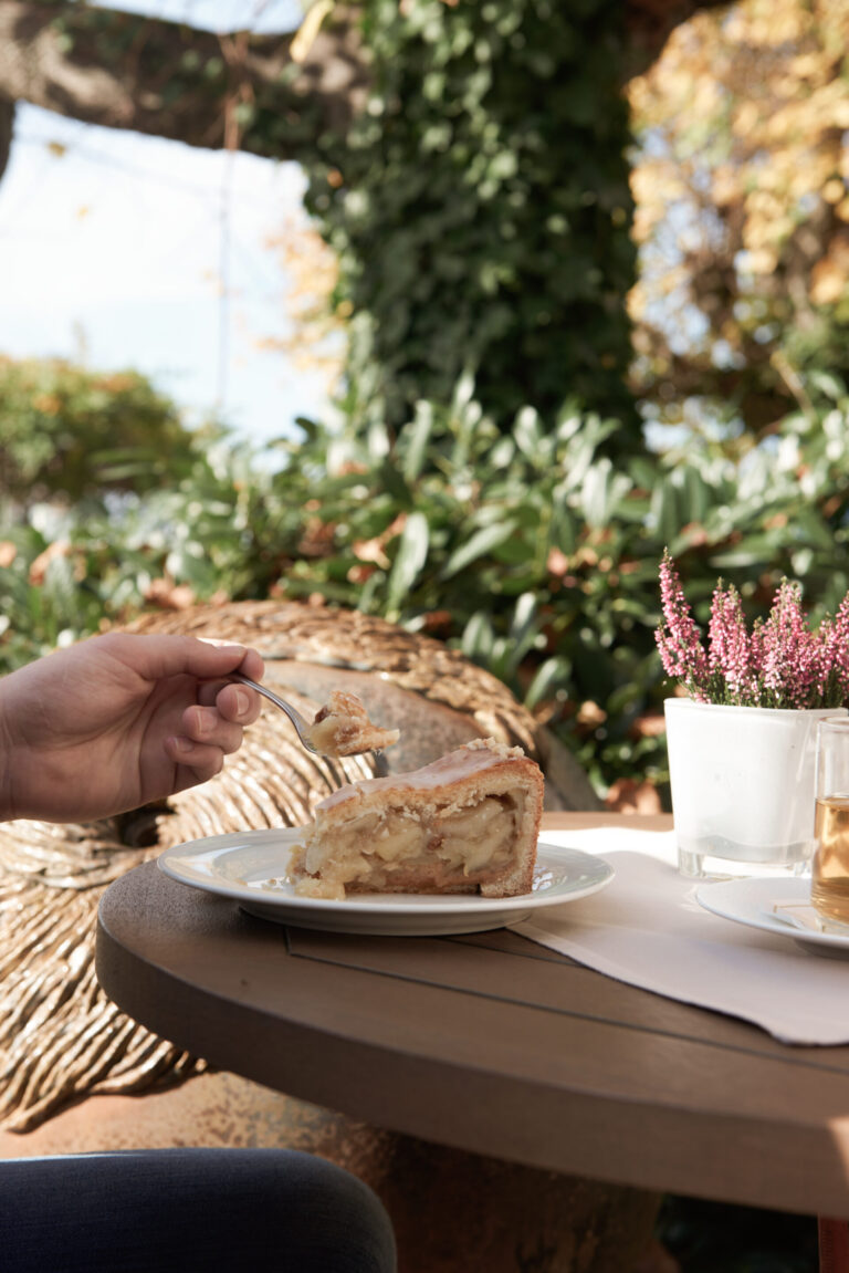 Eine Person hält eine Gabel mit einem Bissen Apfelkuchen an einem Tisch im Freien, umgeben von Grün und einer Topfpflanze.