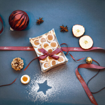 Eine Geschenkschachtel mit Keksen, zusammengebunden mit einer roten Schleife, umgeben von Weihnachtsdekorationen, getrockneten Apfelscheiben und Puderzucker.
