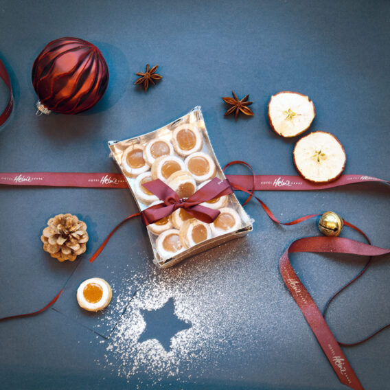 Eine Geschenkschachtel mit Keksen, zusammengebunden mit einer roten Schleife, umgeben von Weihnachtsdekorationen, getrockneten Apfelscheiben und Puderzucker.