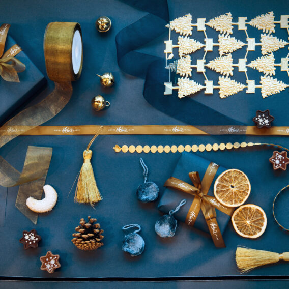 Flache Anordnung von goldenen und blauen Weihnachtsdekorationen, Bändern, Tannenzapfen, getrockneten Orangenscheiben, Ornamenten und verschiedenen festlichen Leckereien.