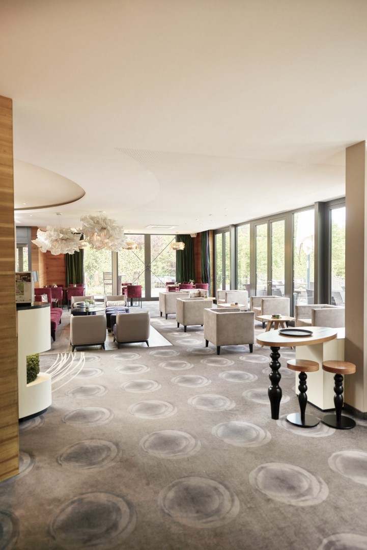 Moderner Loungebereich mit Sesseln, runden Tischen, großen Fenstern und sanftem Tageslicht, der eine helle und einladende Atmosphäre schafft.