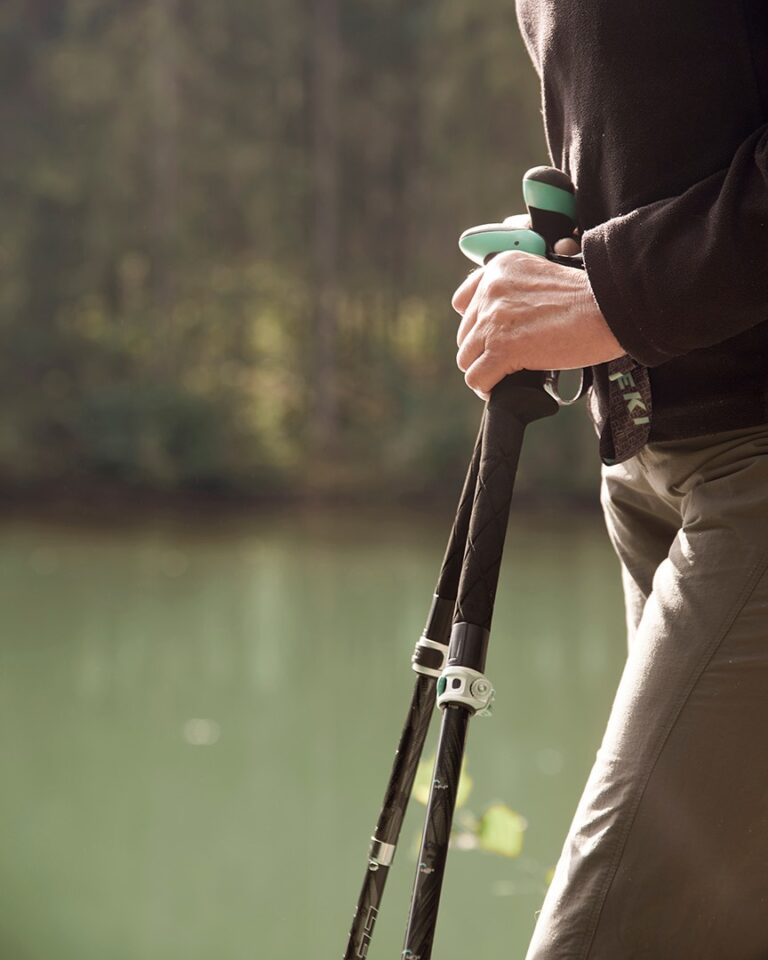 Eine Person steht mit Trekkingstöcken in der Hand an einem See, im Hintergrund sind Bäume unscharf zu sehen.