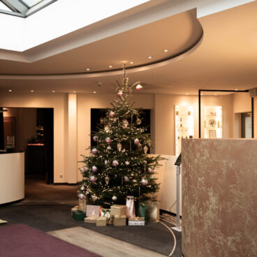 In einem modernen, sanft beleuchteten Lobbybereich steht ein geschmückter Weihnachtsbaum mit verpackten Geschenken darunter.